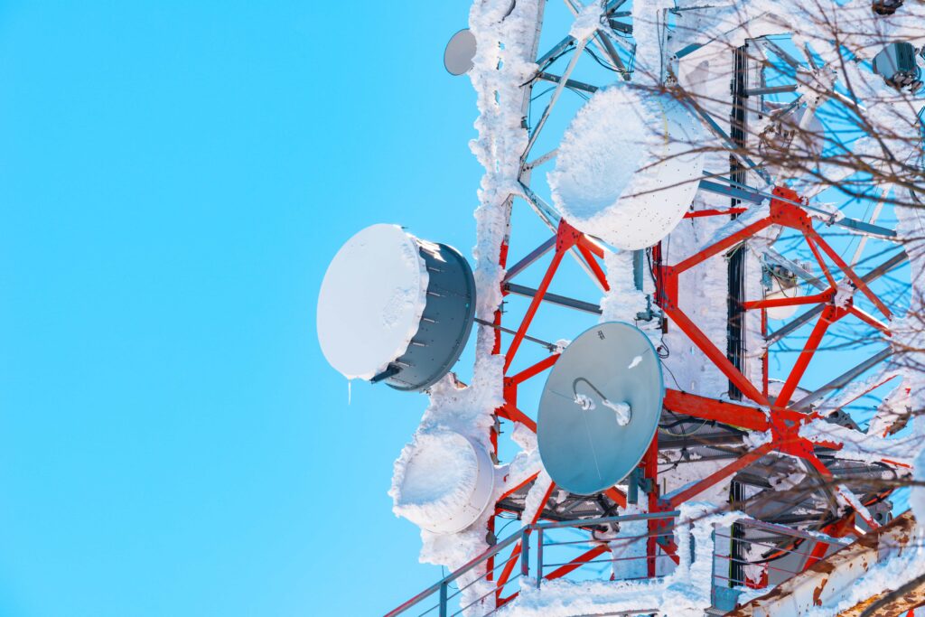 A telecommunications tower with multiple satellite dishes and antennas attached to its red and white metal framework, set against a bright blue sky.