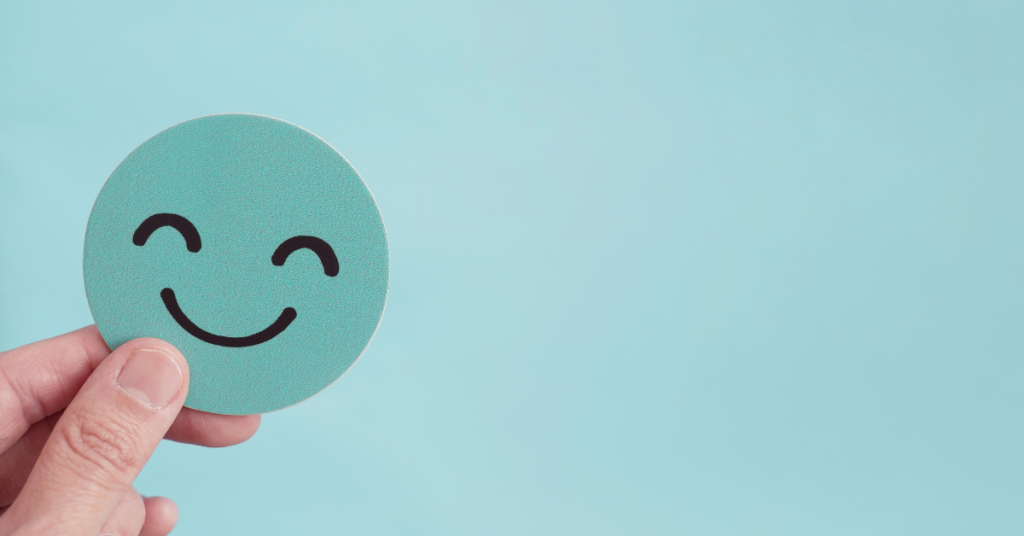 A hand holding a round teal card with a black smiley face on it, set against a solid light blue background.