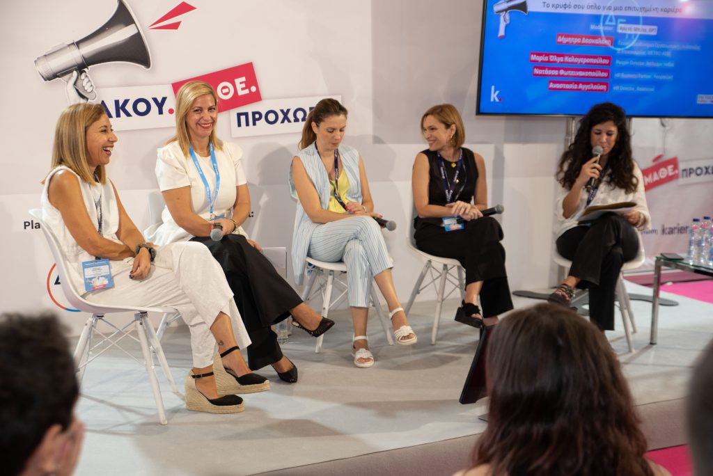 A panel discussion at an event with five speakers seated on white chairs, holding microphones.